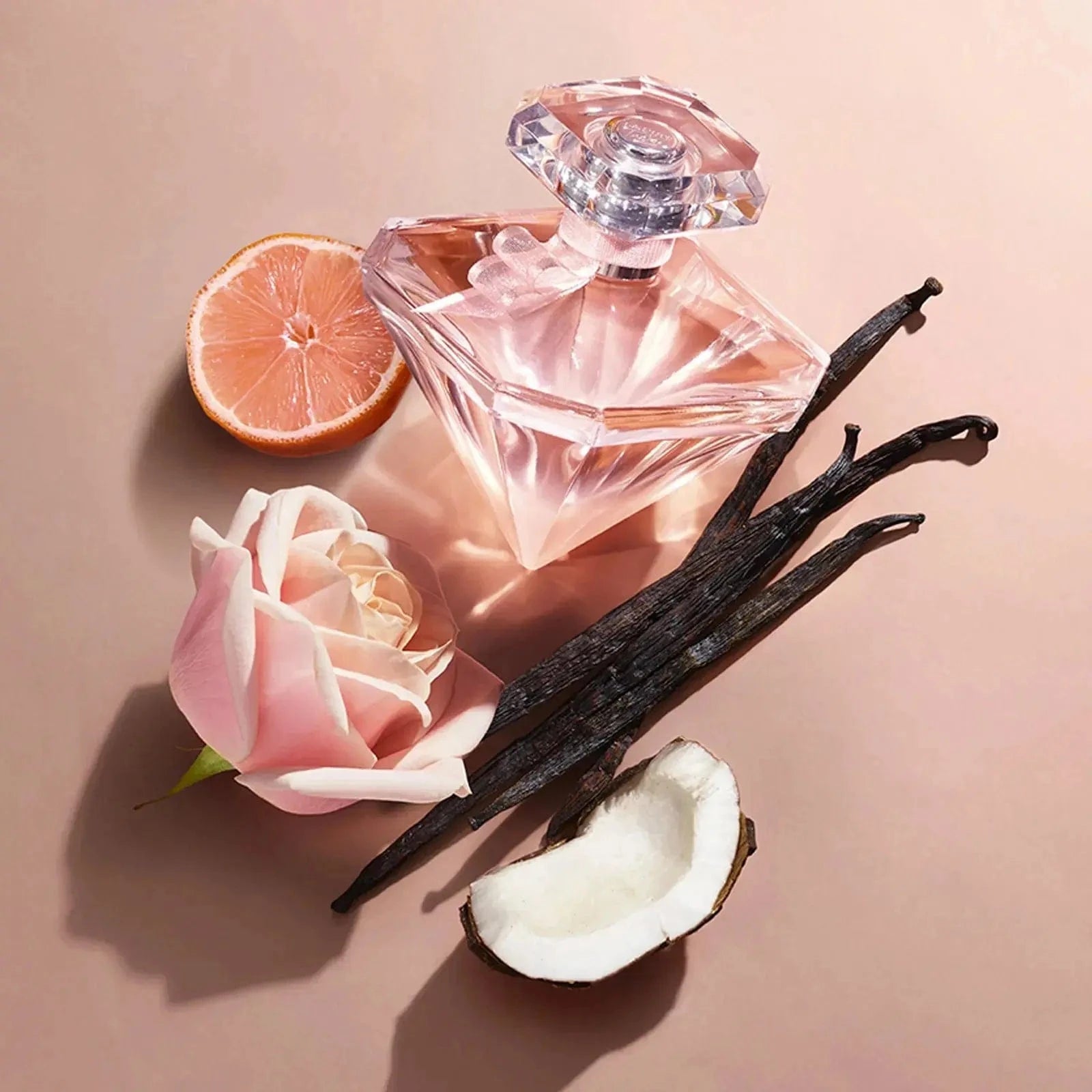 An artistic flatlay of the La Nuit Trésor Nude bottle, surrounded by its key ingredients: a pink rose, vanilla beans, a piece of coconut, and a slice of bergamot.
