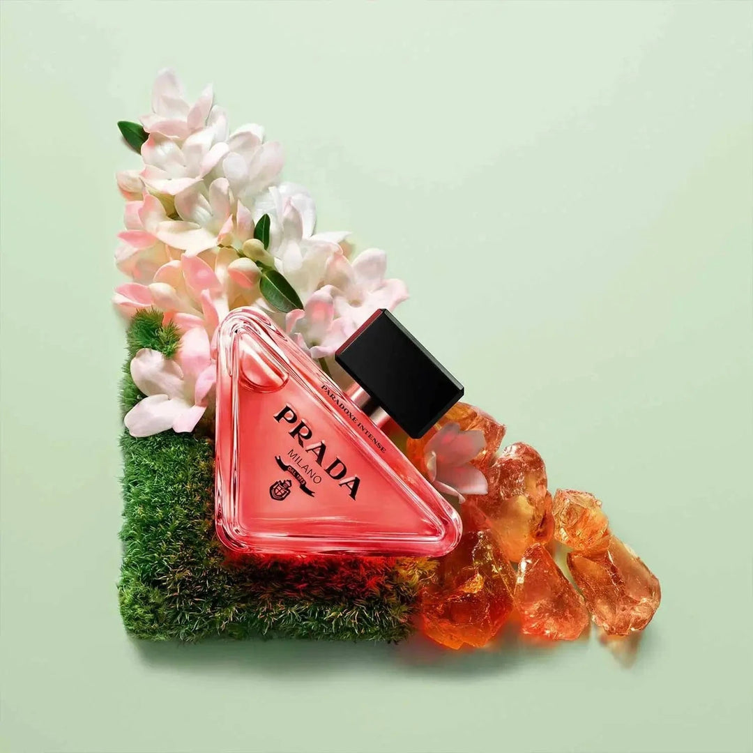 An artistic flatlay of the Prada Paradoxe Intense bottle, resting on green moss with white flowers and amber-colored gems.