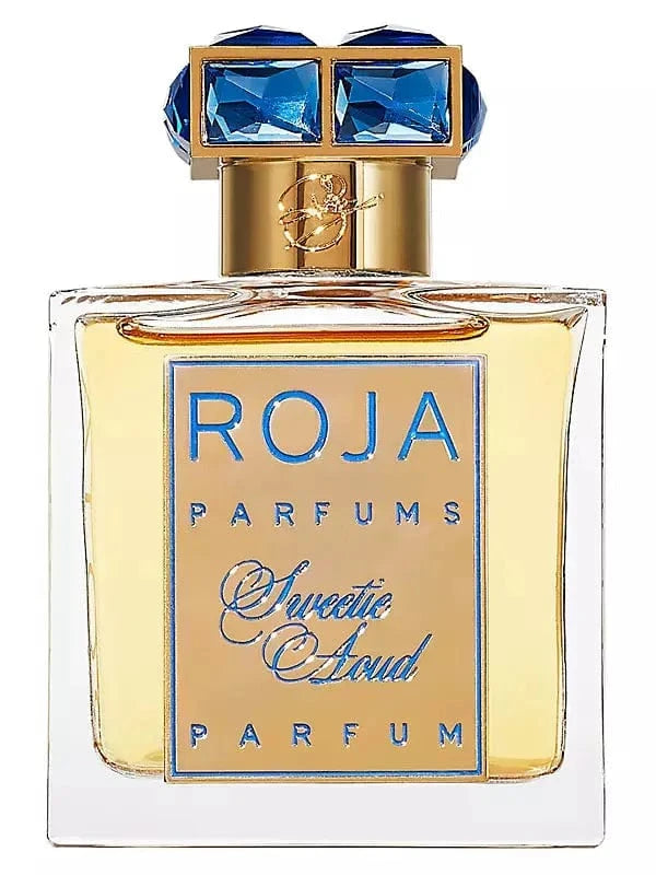 The luxurious Sweetie Aoud by Roja Parfums bottle with its gold label and blue crystal-embellished cap, shown against a softly blurred, opulent room.