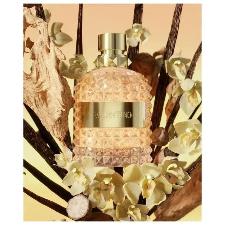 A stylized photo of the Valentino Uomo Born in Roma The Gold bottle, surrounded by vanilla orchids and dried wood pieces.